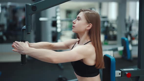 A beautiful girl with her strong arms performs an exercise on a simulator alt
