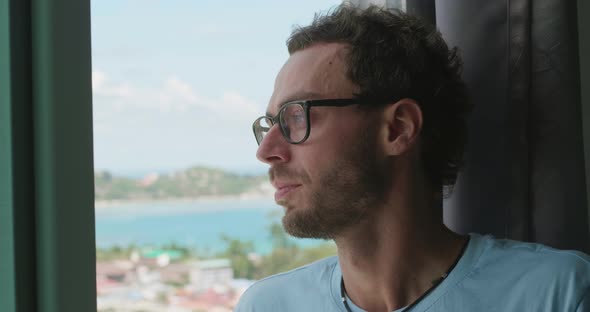 Relaxed Caucasian Man in Casual Blue Tshirt Putting on Glasses for Vision and Gazing Out the Window alt