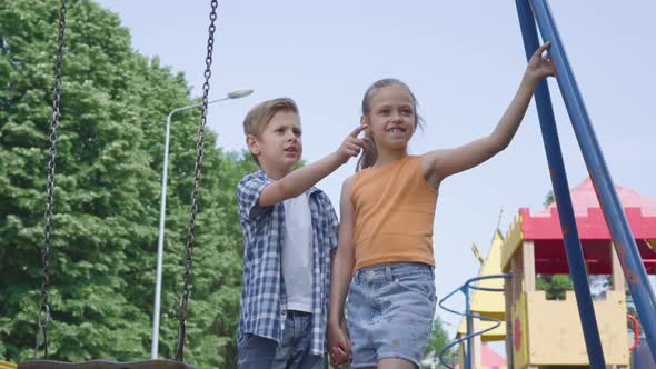 Cute Boy and Beautiful Girl with Long Hair Pointing Away Holding Hands Near the Swing, Smiling alt