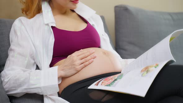 Asian young woman pregnant sitting and reading book with smile on sofa. alt