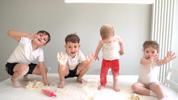Four Small Children of Different Ages Sit on a Table in the Kitchen and Wave Their Hands at the