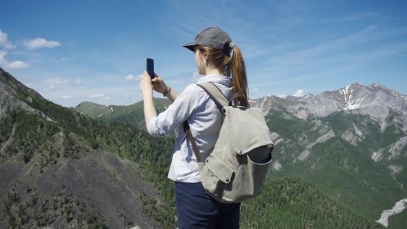 Woman Traveler in a Cap with a Hiking Backpack Takes Pictures of the Mountains Using the Phone