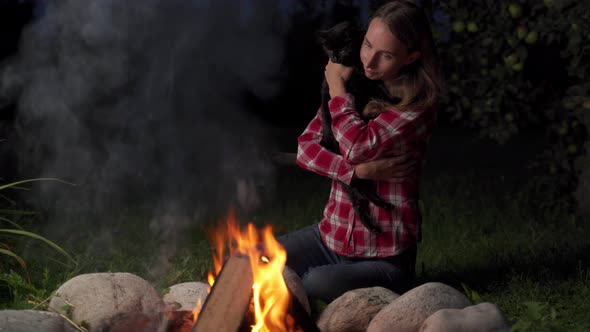 Woman Holds a Cat and Sits Near Campfire at Evening alt