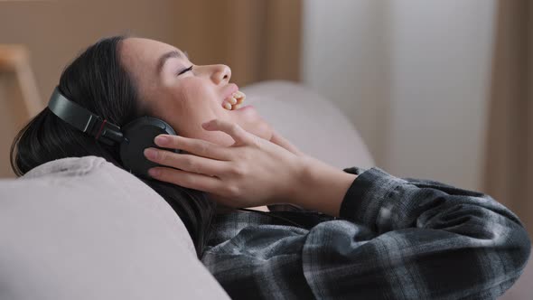 Calm Asian Korean Millennial Woman in Headphones Sitting Lying on Comfortable Home Sofa Couch with alt