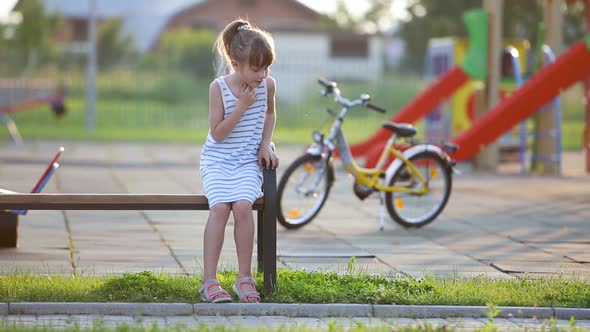 Cute Little Girl Sitting on a Bench and Riding a Bicycle on a School Yard in Summer alt