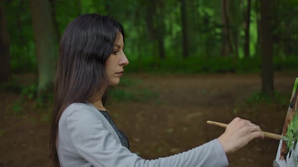 Side View of a Girl Painting a Landscape in a Forest. alt