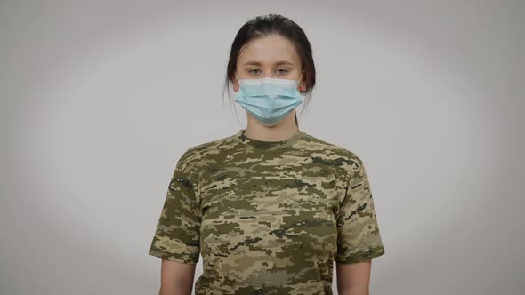 Front View Portrait of Serious Young Military Woman in Coronavirus Face Mask Posing at Grey alt