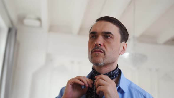 Man in a Blue Shirt Ties Tie with His Hands Near a Mirror in a Large White Room alt