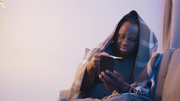 Ill African American Black Man Covered in Blanket Checking Temperature and Using Phone alt