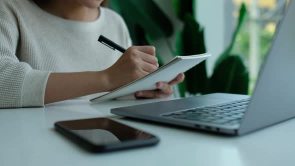 Closeup of a woman writing on a notebook with laptop computer and smartphone on the table