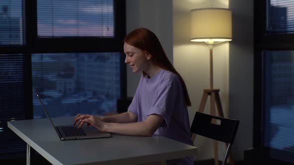 Cheerful Redhead Young Woman Freelancer Using Laptop Computer for Work Sitting at Desk Near Window alt