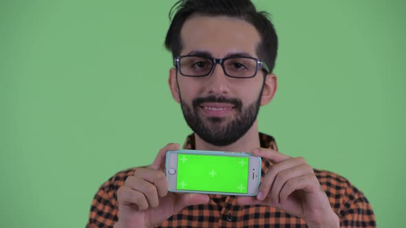 Face of Happy Young Bearded Persian Hipster Man Thinking While Showing Phone alt