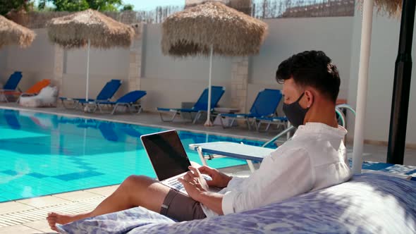 Man Works in Mask Using Laptop By Swimming Pool in Hotel at COVID-19 Pandemic alt