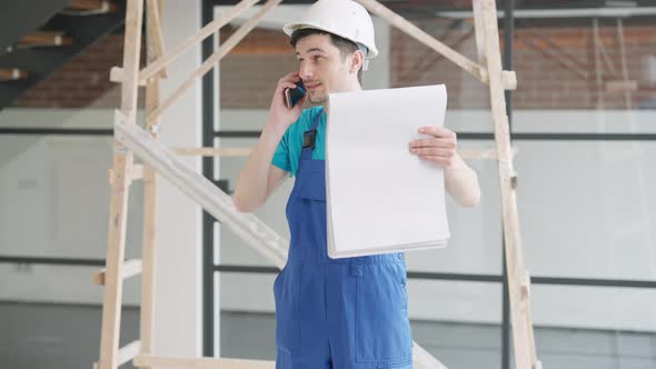 Confident Young Man in Hard Hat Talking on the Phone Holding Drawings alt