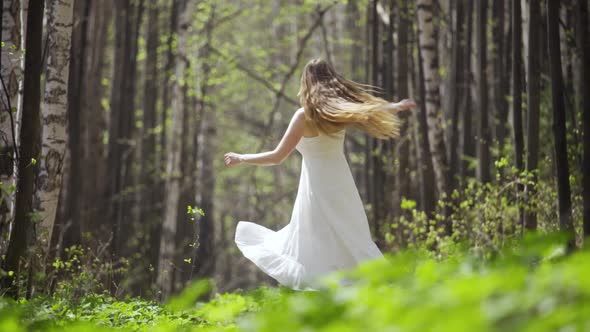 A Cute Young Woman Whirls Around Herself in a White Dress in a Light Forest alt