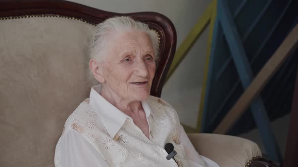 Smiling Grandmother Sitting on Sofa in Room and Talking with Microphone alt