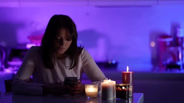 Young Woman is Using Social Nets in Smartphone Sitting on Kitchen at Night Candles are Flaming on alt