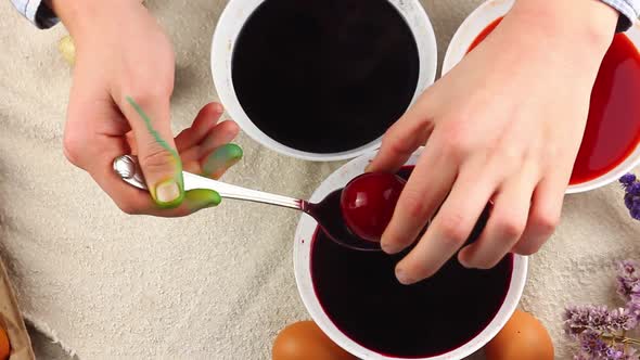 Caucasian Child Paints an Easter Egg with a Metal Spoon and Dips It Into a White Bowl with Red Dye alt