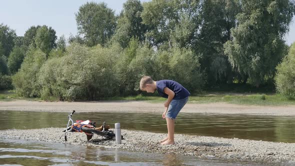 Cute Caucasian Teenager Barefoot Throws Stones Into the Water While Standing on the Bank of the alt