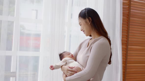 Beautiful Asian woman holding newborn baby in her arms standing in front of windows alt