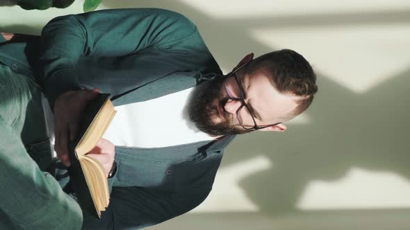 Bearded Young Man Sitting Near Window and Reading a Book Vertical Video