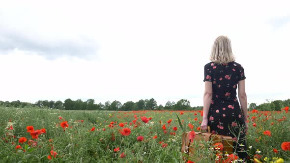 Blonde girl in beautiful dress with suitcase in poppies field in summer time