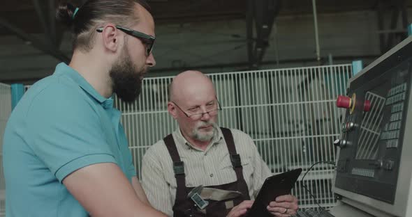 Two Engineers Work with Control Panel of Machine, Use Tablet and Discuss alt