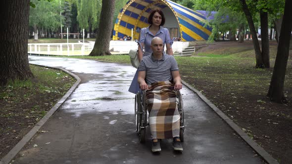 A Woman Is Driving a Young Man with Oncology in a Wheelchair Through the Park. The Man Is Bald Due alt