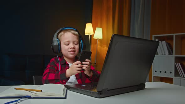 Cheerful Energetic Little Boy Using Smartphone and Laptop and Listening to Music in Headphones at alt