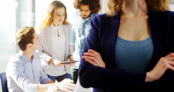 Woman standing with arms crossed while colleague discussing in background alt