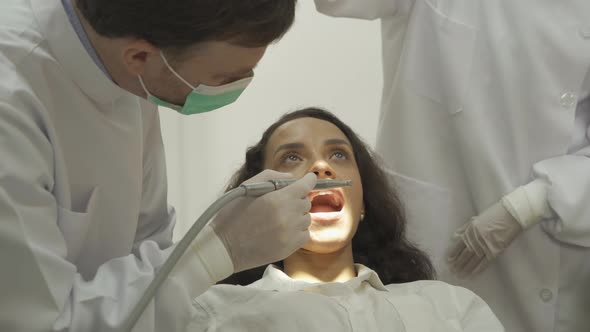 A dentist checking up Latin woman's teeth by using dental mouth mirror at hospital clinic alt