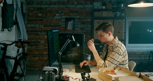 Man Works on Computer Eating Pizza in Loft Electronics Workshop or Home Garage
