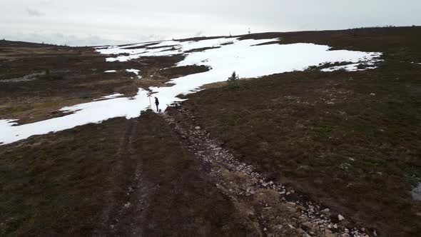 A woman and her french bulldog walking on a mountain gravel path with snow infront of them. Aerial f alt