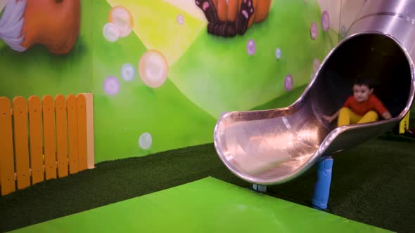 Little Brunette Boy Rolls Down a Slide in an Amusement Park