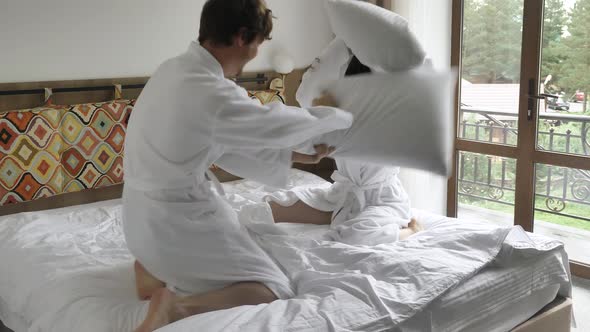 A Cheerful and Active Married Couple in White Coats Fights with Pillows on a Soft Bed in a Hotel alt