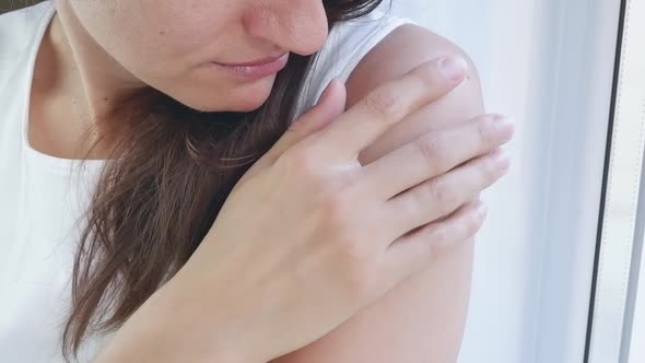 A Woman in a White Tshirt By the Window Touches a Large Bruise on Her Arm with Her Hand alt