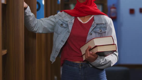 Asian female student wearing a red hijab selecting a book from a bookshelf alt