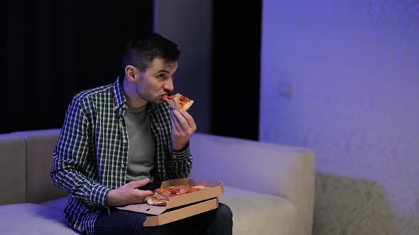 Handsome Young Man Eating Pizza While Watching TV While Relaxing at Sofa in the Living Room