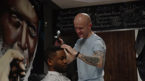 A Bald Hairdresser Dries the Head of a Black Barbershop Client with a Hairdryer alt