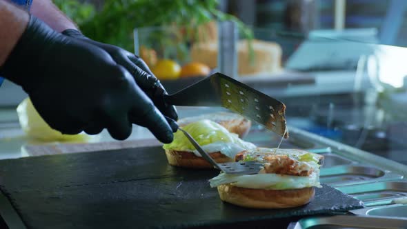 Male Chef in Black Gloves Cooks Juicy Tasty Burgers with Cheese and Meat on a Cutting Board alt
