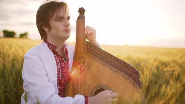 Young Handsome Bandurist Folk Ukrainian Musician Playing on Bandura at Sunset in Wheat Field