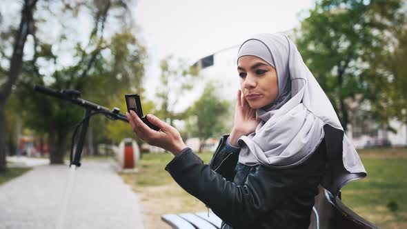 Portrait of Young Muslim Woman Looking at Herself Using Hand Mirror Outdoors in Park alt