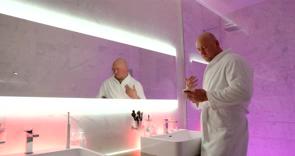 Hilarious Bald Man in the Bathroom in the Evening Dressed in a Robe Stands in Front of a Mirror with alt