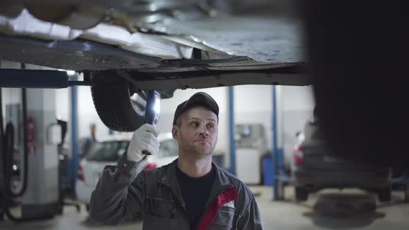 Serious Caucasian Man in Workwear and Cap Walking Under Car Bottom with Flashlight. Male Adult Auto alt
