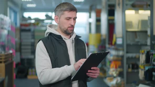 Concentrated Hardware Store Worker Standing with List of Products Checking Goods on Shelves alt