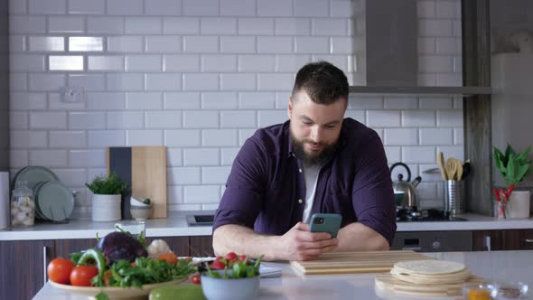 Handsome Young Man with a Smartphone in the Kitchen using Food App and Smiling alt