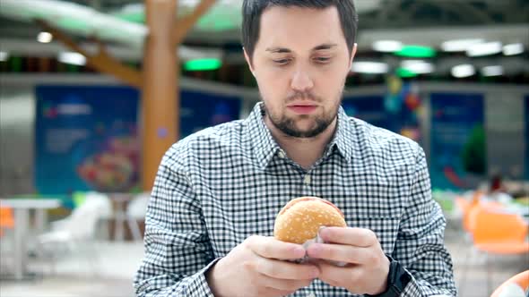 A Young Man Is Eating a Hamburger with Meat in a Food Court. alt