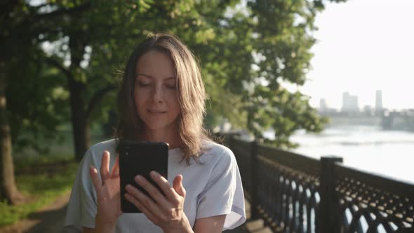 a Woman Walks and Looks Carefully at the Phone Screen on the City Embankment alt