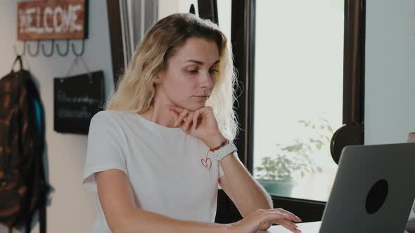 Serious blonde woman in headphones sitting in cafe with laptop making video calling alt