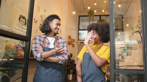 Two baristas at a cafe door, arms crossed, teasing together with a happy smile. alt